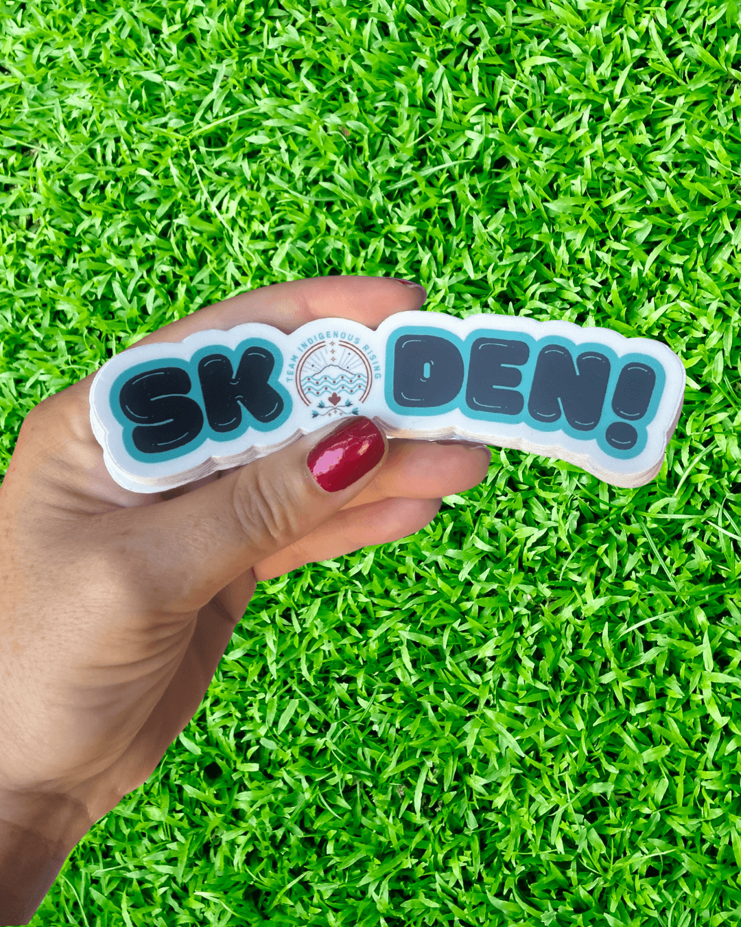 Hand holding a stack of 'SKODEN!' stickers against a grass background