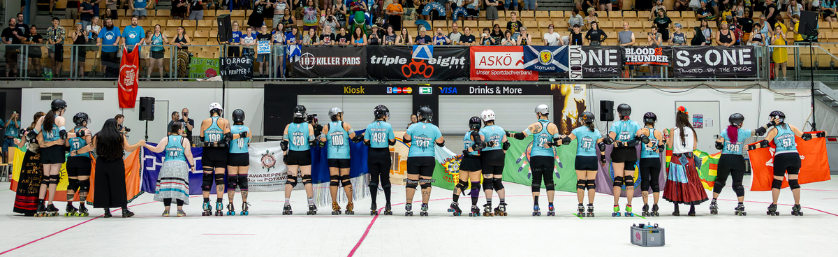 Indigenous Rising Roller Derby teammates stand with their backs facing the camera holding their national/tribal flags.