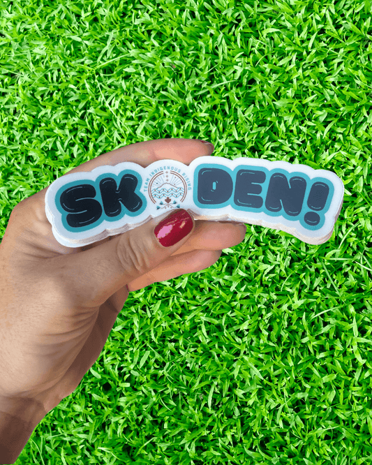 Hand holding a stack of 'SKODEN!' stickers against a grass background