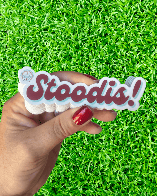 Hand holding a stack of 'Stoodis!' stickers against a green grass background